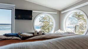 A bedroom with a bed, a TV mounted on the wall, and two large windows offering a view of the sea. - Waterfront Escape at the Tip of Door County (Ellison Bay)