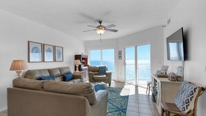 A spacious living room with a large sofa, a smaller sofa, a coffee table, and a TV mounted on the wall. There is a ceiling fan, a window with a view of the ocean, and a wooden chair.