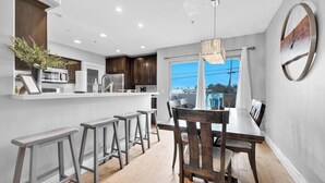 A modern kitchen with a dining area, wooden bar stools, and a large window offering a view.