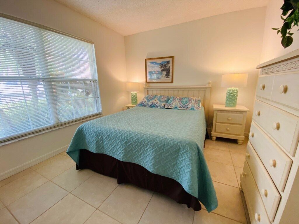 Une chambre à coucher comprenant un lit, des tables de chevet, une commode et une fenêtre avec des stores.
