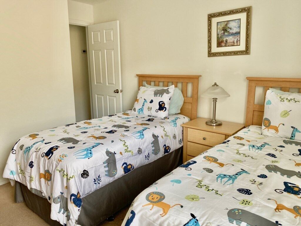 A bedroom with two beds, each with a white bedspread featuring animal prints. There is a wooden headboard, a bedside table with a lamp, and a door leading to another room.