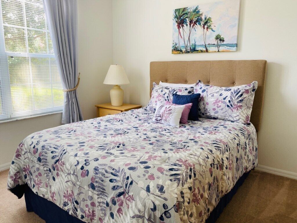 Une chambre à coucher avec un lit, une table de chevet, une lampe, une fenêtre avec des rideaux et un tableau au mur.