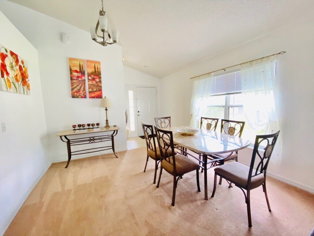 A dining area with a round table and six chairs, a bench, a chandelier, and two paintings on the wall.
