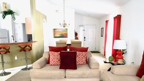 A living room with a beige sofa, red cushions, a green rug, a dining area with a bar, and a red lamp. - Gated Community - Southern Dunes Golf & Country Club - Private Pool (Haines City)