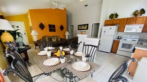 Un comedor con una mesa de vidrio y sillas de metal, una cocina con electrodomésticos blancos y una pared de color naranja brillante.