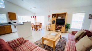 A living room with a sofa, a coffee table, and a TV stand. There is a kitchen area with cabinets and a sink. - Golf Course Views - Heatable Pool -  Southern Dunes (Haines City)