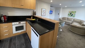 Una cocina moderna con armarios de madera, encimera negra y lavavajillas. En la sala de estar, hay un sofá color crema y un cuadro.