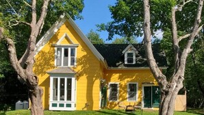Une maison jaune avec un toit à pignon, de grandes fenêtres et un porche à l’avant.