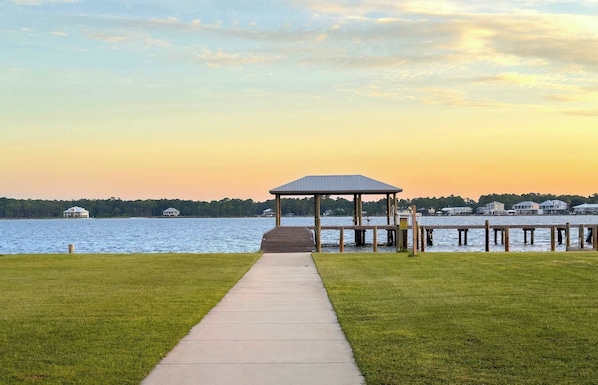 A gazebo with a wooden deck leading to a lake, surrounded by grassy areas and a dock extending into the water.