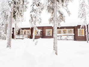A wooden cabin with a snow-covered roof and windows, surrounded by snow-laden trees.