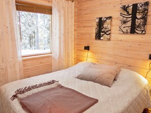 A wooden cabin bedroom with a bed, two pillows, a blanket, a window with curtains, and two wall-mounted light switches. - Villa jaloranta a by Interhome (Kuusamo)