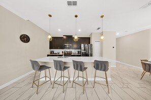 Une cuisine moderne avec un îlot central, des tabourets de bar, des luminaires suspendus et une horloge au mur.