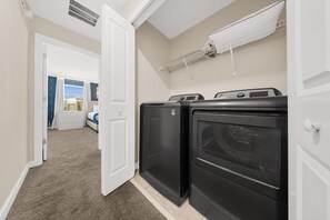 Une buanderie équipée de deux lave-linge et deux sèche-linge à chargement frontal, une porte blanche donnant sur une chambre, et une étagère blanche au-dessus des sèche-linge.