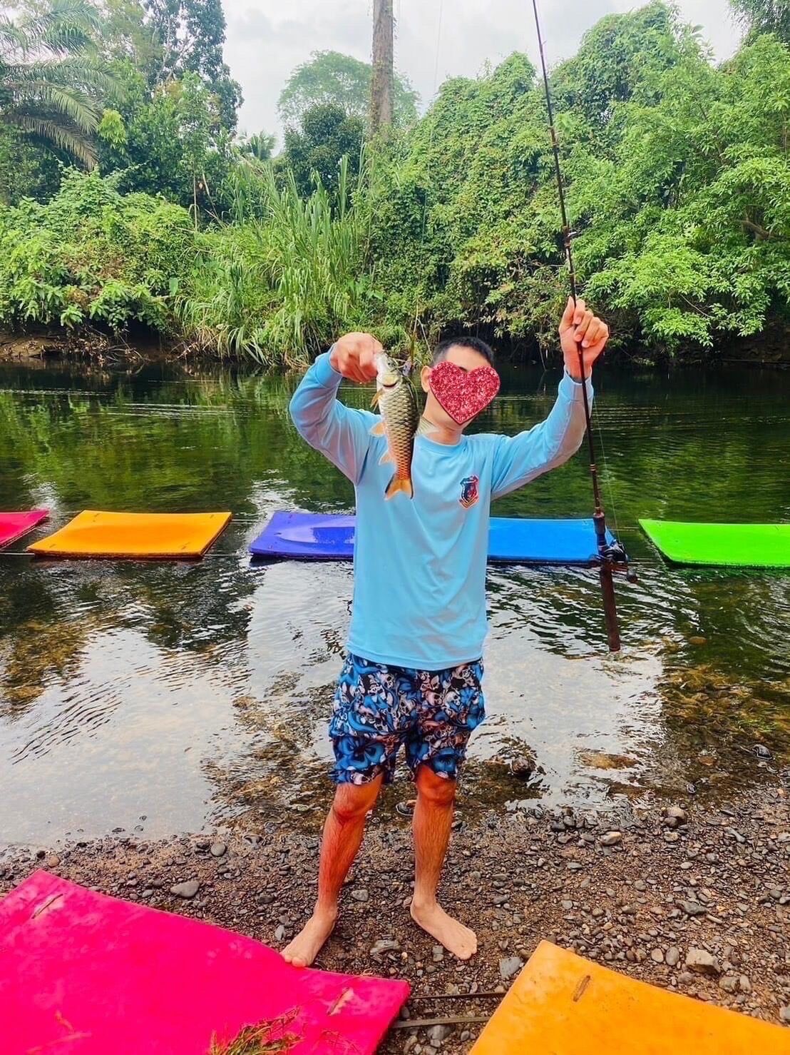 A person standing on a rocky shore holding a fish, with colorful mats and a river in the background.