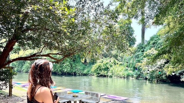 Una mujer parada junto a un río, con una mesa y sillas, contemplando el río rodeado de vegetación.
