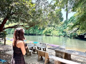 Una mujer parada junto a un río, con una mesa y sillas, contemplando el río rodeado de vegetación.