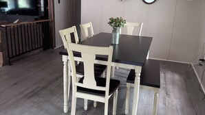 Zona pranzo con un tavolo in legno scuro e sedie bianche, un vaso con fiori, uno specchio e una ghirlanda appesa al muro.