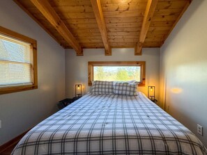 A bedroom with a wooden ceiling, a bed with plaid bedding, two bedside lamps, and a window with blinds.