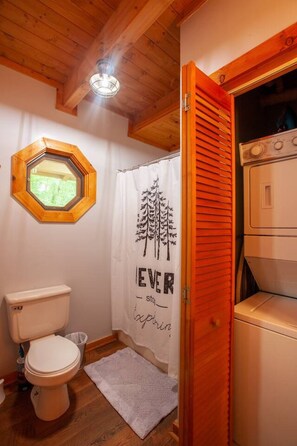 A small bathroom with a wooden ceiling, a hexagonal window, a toilet, a shower curtain with a tree design, and a washing machine. - Almost Heaven - Minutes to the Parkway!! (Pigeon Forge)