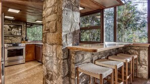 Una cucina con pareti in pietra, un angolo bar con sgabelli e un'ampia finestra che si affaccia su alberi.