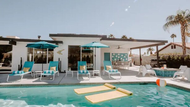 Zona de piscina con sillones de descanso y sombrillas, una mesa junto a la piscina y vistas a un edificio moderno.