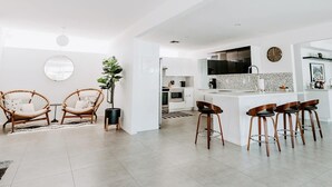 Una cocina moderna con armarios blancos, una isla central con taburetes y un comedor con sillas de mimbre.