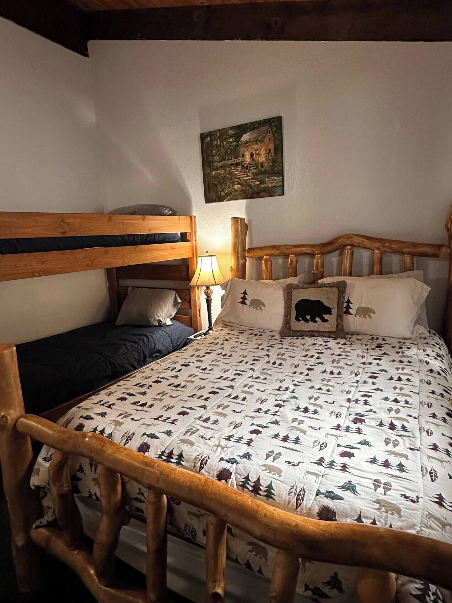 Una habitación con una cama nido de madera, una cama individual con colcha estampada, una lámpara de noche y un cuadro en la pared.
