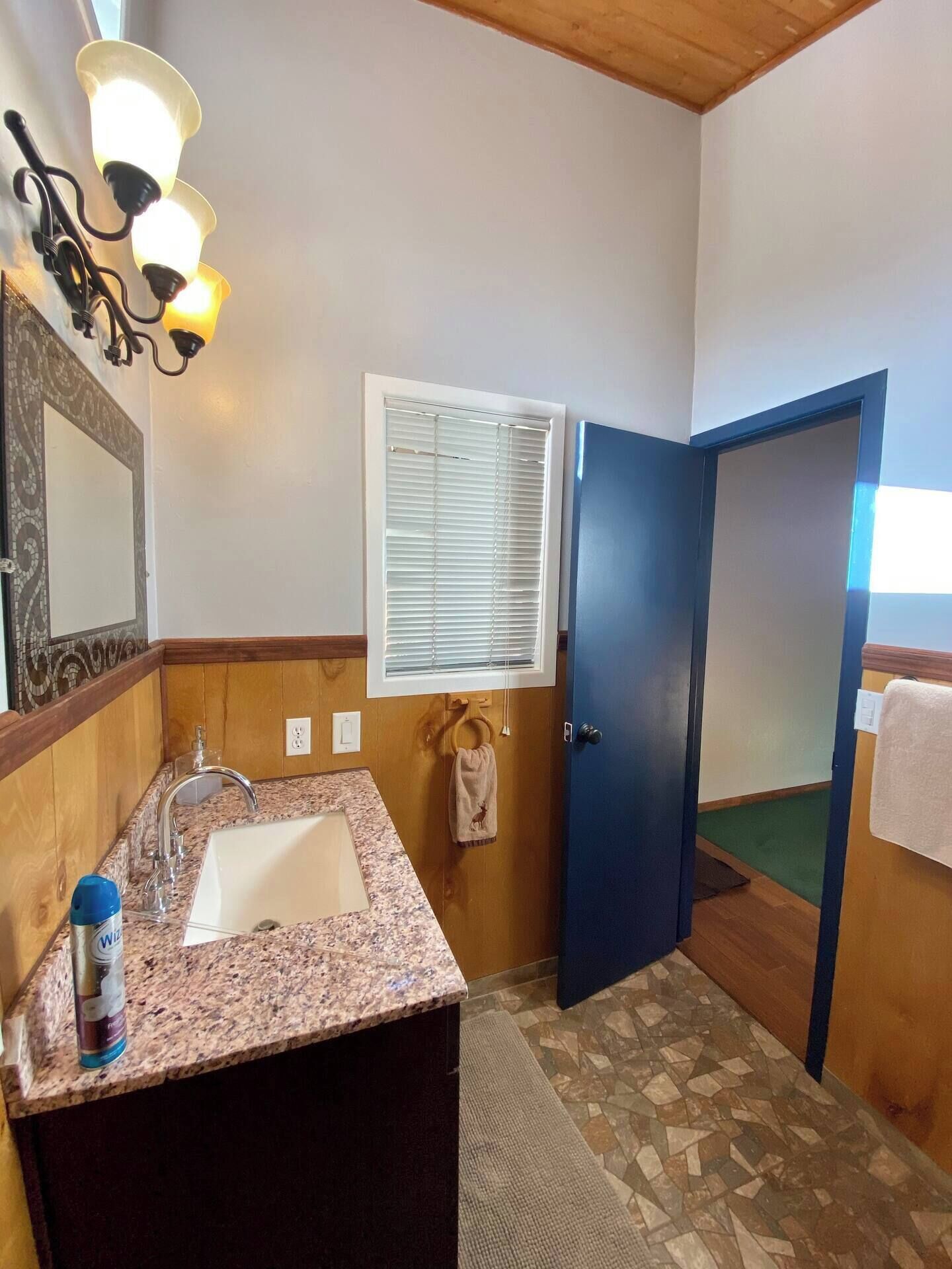 Un baño con lavabo de encimera de granito, un espejo con marco decorativo, un perchero con toalla, una ventana con persianas y una puerta.