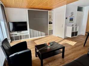 A modern living room with a flat-screen TV, a black coffee table with fruit, and a wooden ceiling.