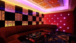 Une pièce faiblement éclairée, avec un coin salon doté de sièges rembourrés rouges, un tapis à motifs et un plafond orné de luminaires aux formes géométriques.