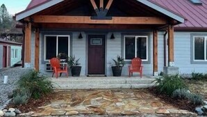 Une maison à deux étages avec un toit rouge, un porche à l’avant orné de deux chaises rouges et un chemin de pierre menant à l’entrée.
