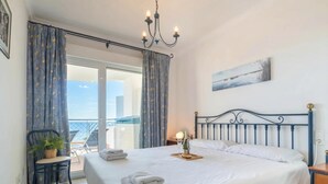 A hotel room with a large bed, a chandelier, a balcony with a chair and table, and a painting on the wall.