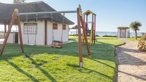 A playground with swings and a swing set, a building with a thatched roof, and a stone pathway.