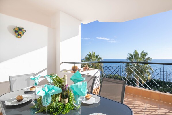 A balcony with a table set for two, featuring a bottle of champagne, a glass, and a cupcake. The balcony overlooks a coastal view with palm trees and the ocean.