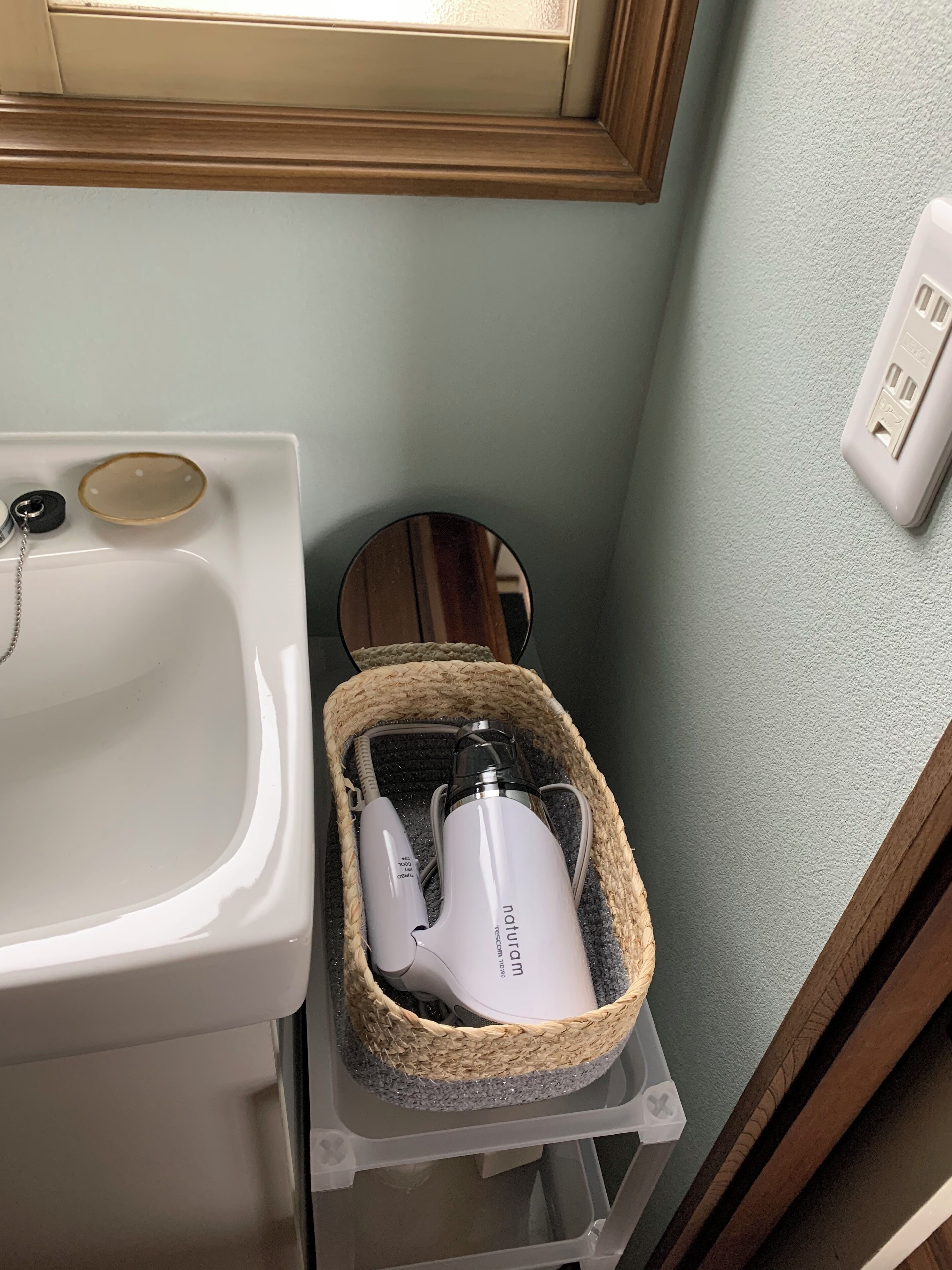 A bathroom with a white sink, a mirror, a white electric toothbrush in a woven basket, and a power outlet on the wall.