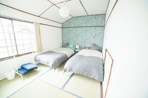 Una habitación de estilo japonés tradicional con suelo de tatami, dos camas con ropa de cama gris, una lámpara colgante y una ventana con cortina.