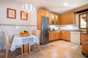 Une cuisine avec des meubles en bois, un réfrigérateur en acier inoxydable, une table à manger agrémentée d’un panier de fruits et un lustre.
