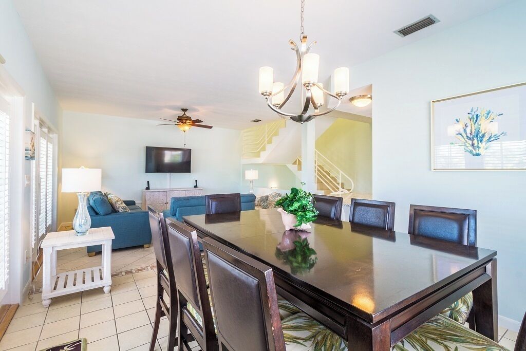 A dining area with a large table, chairs, a chandelier, a TV, and a staircase in the background.