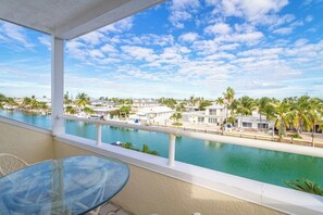 A balcony with a glass table and chairs overlooking a swimming pool and a resort area with palm trees and buildings.
