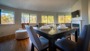 A dining area with a wooden table set for four, surrounded by chairs, large windows with autumn foliage, and a fireplace. - Tree House in Center of Helen (Helen)