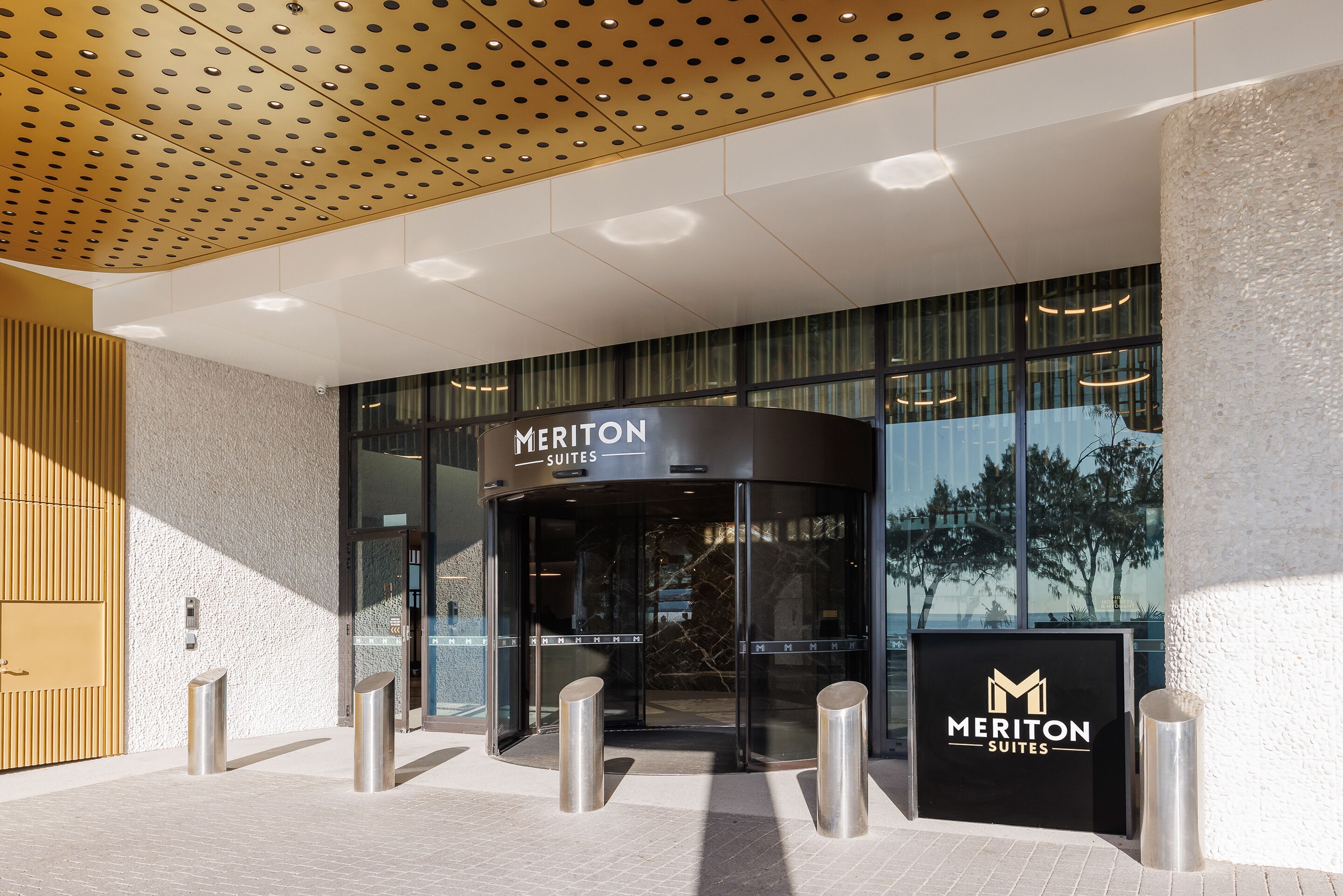 A hotel entrance with a revolving door, a sign reading "Meriton Suites", and a glass facade reflecting trees.