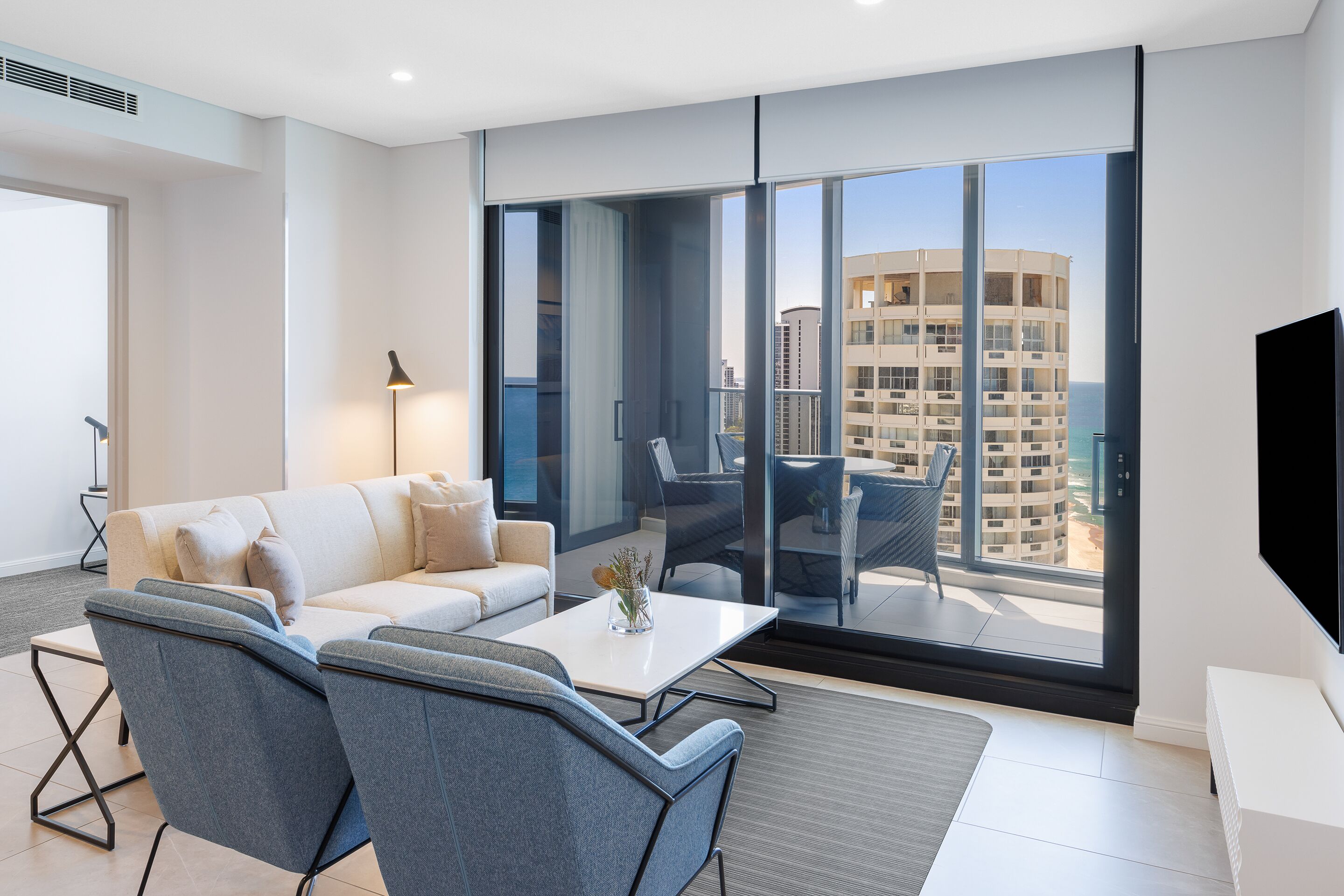 Ein modernes Wohnzimmer mit Sofa, Sesseln, Couchtisch und Fernseher. Große Fenster bieten Blick auf die Stadt.