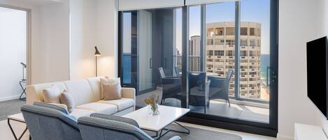 A modern living room with a sofa, chairs, a coffee table, and a TV. It features large windows with a city view.