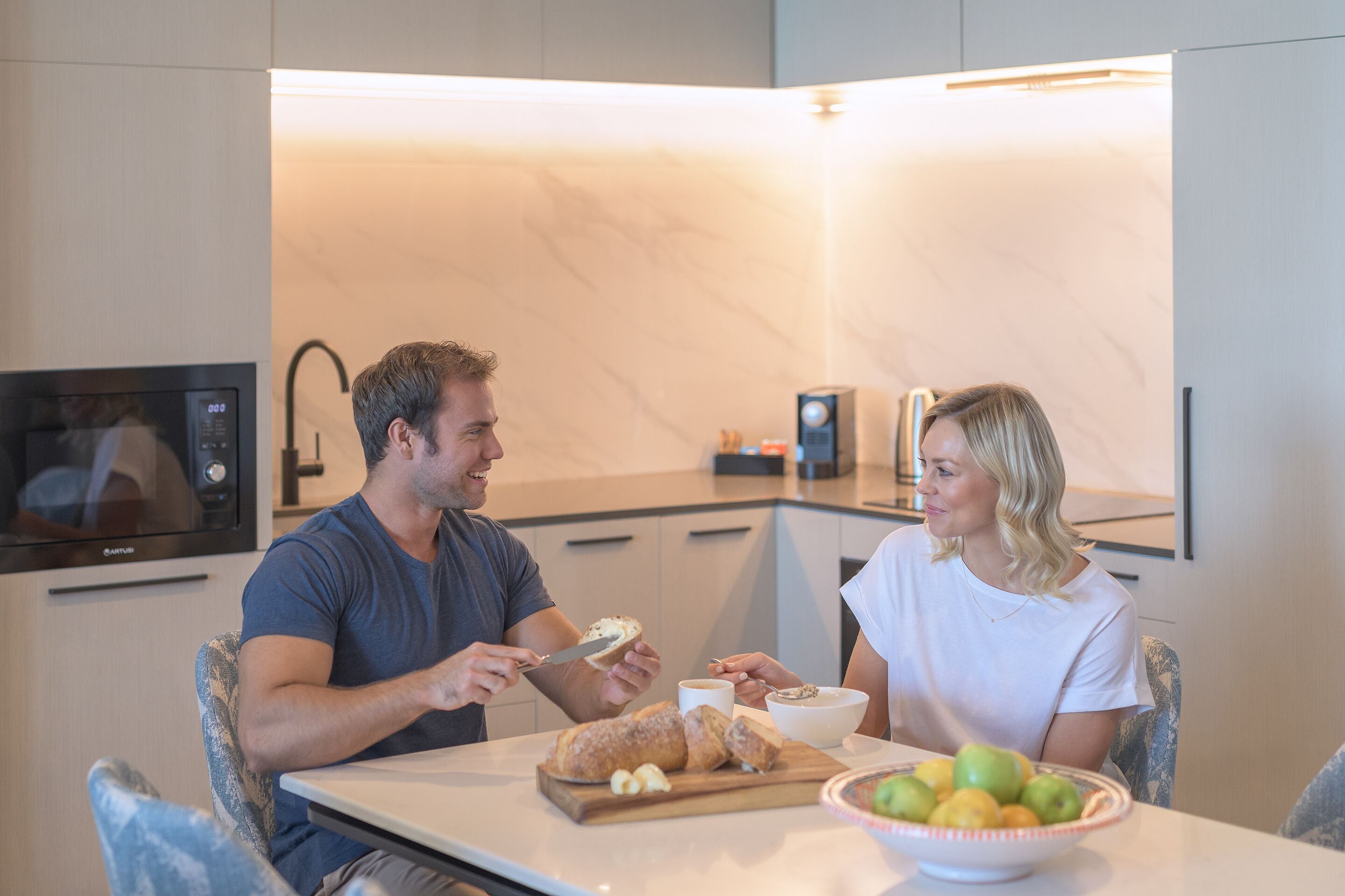 Una cocina moderna con microondas, cafetera y una mesa de comedor con pan y fruta.