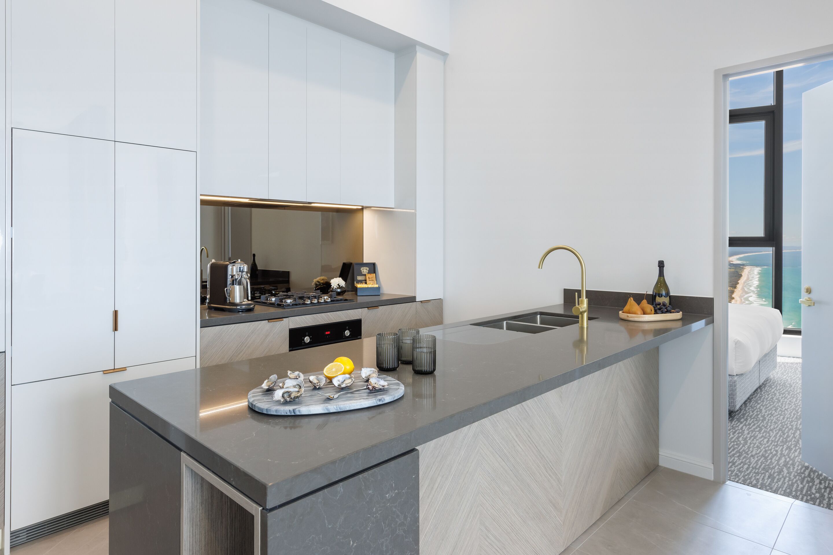 A modern kitchen with a large island, a built-in oven, and a view of the beach through a window.