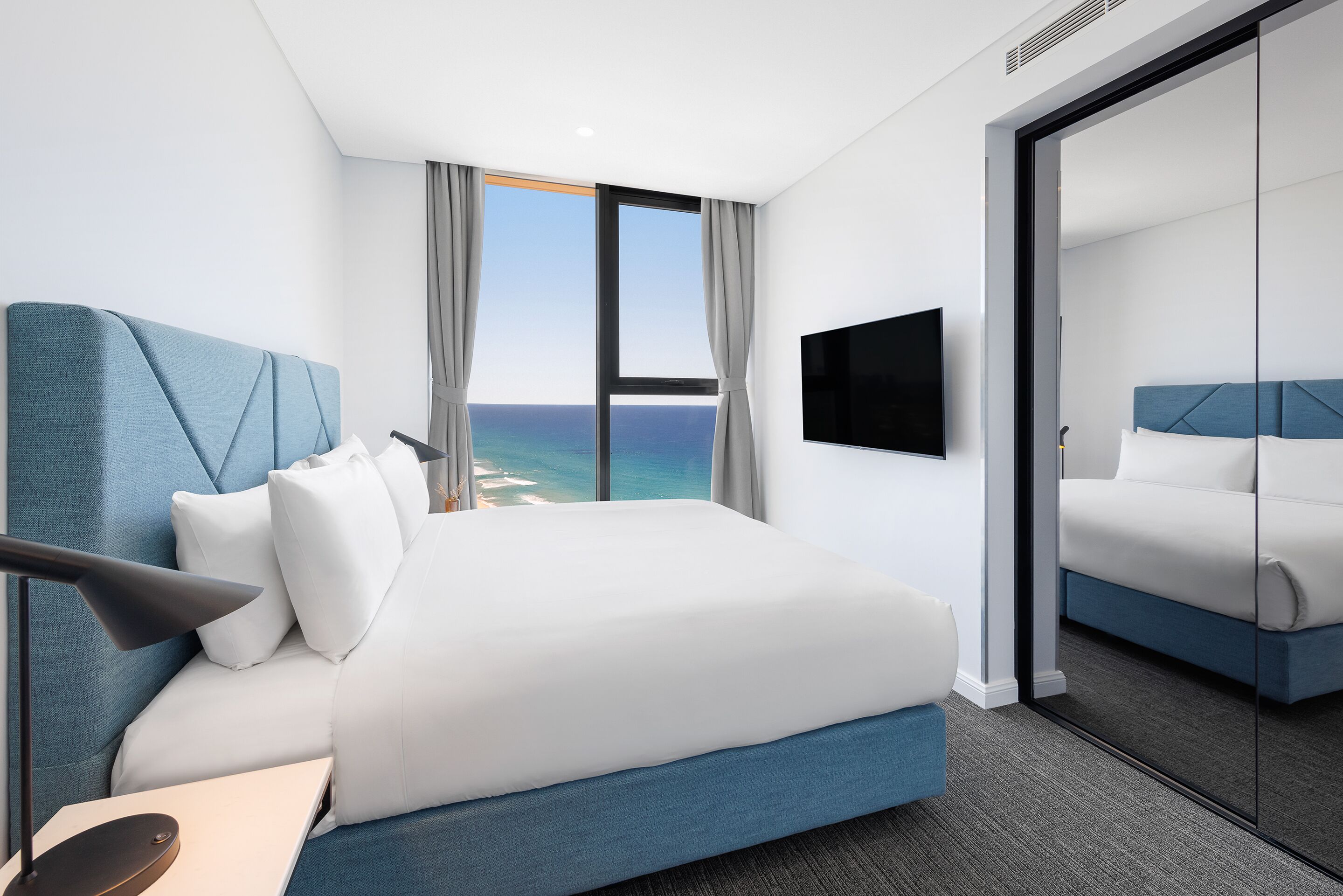 A modern hotel room with a large bed, a flat-screen TV, and a view of the ocean through large windows.