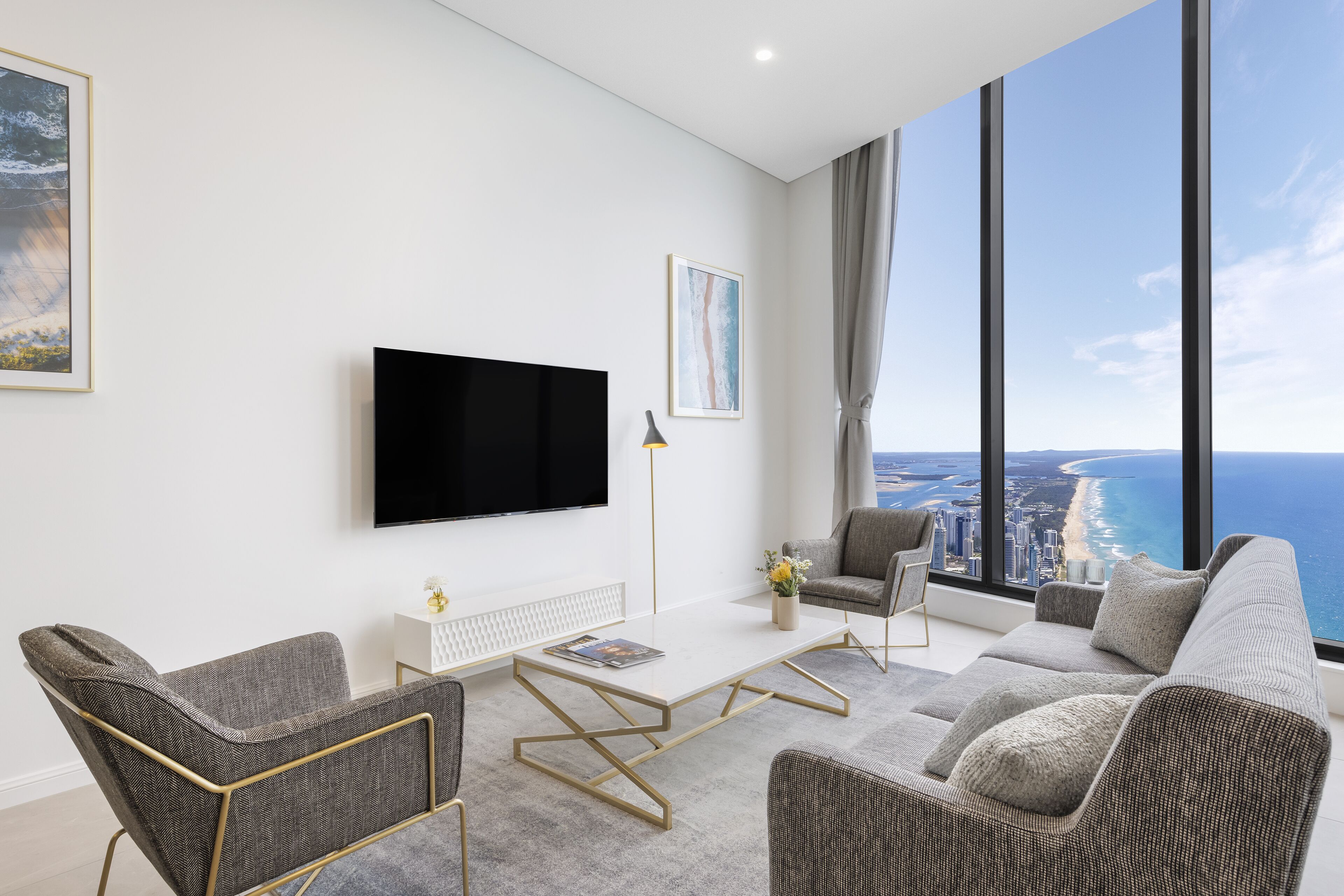 A modern living room with a flat-screen TV, grey upholstered furniture, and a large window offering a view of the coastline.
