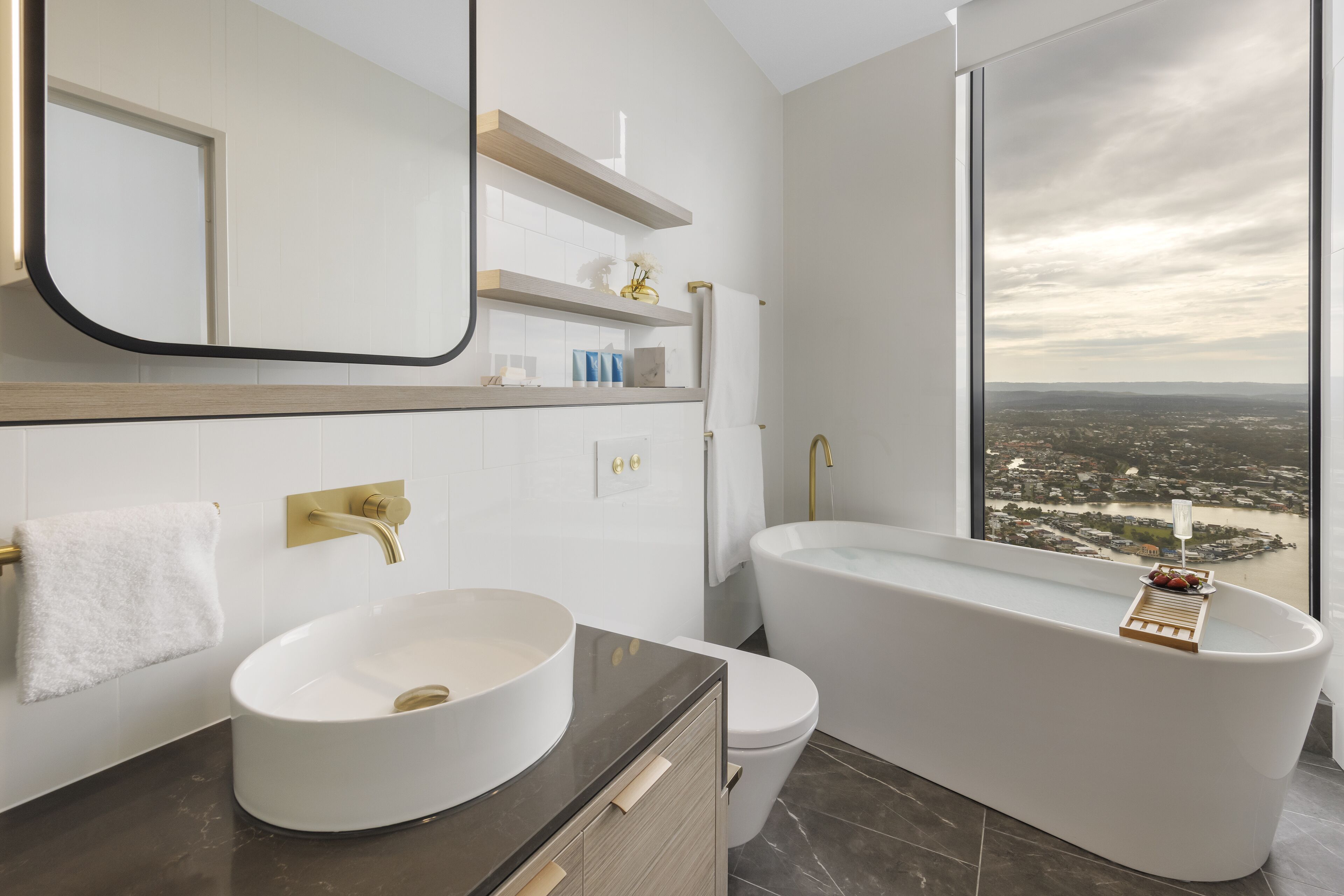 A modern bathroom with a large bathtub, a round sink, and a toilet. There is a large window providing a view of a cityscape.