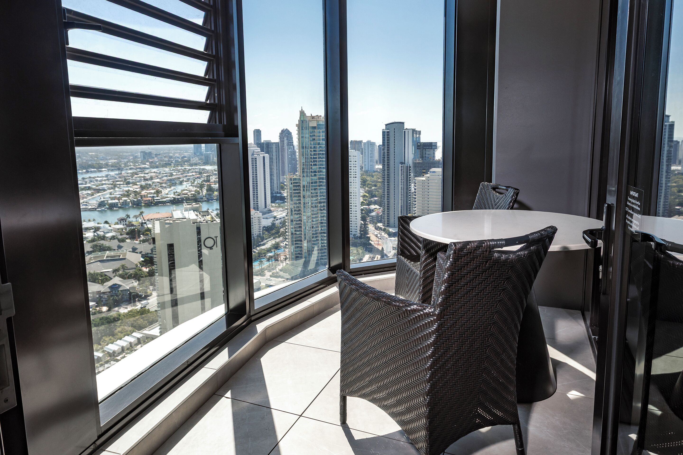 Een balkon van een hoog gebouw met een panoramisch stadsgezicht, een ronde tafel en rieten stoelen.