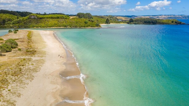 Une vue aérienne d’une plage aux eaux turquoise et limpides, bordée d’un rivage sablonneux, avec des collines verdoyantes en arrière-plan.